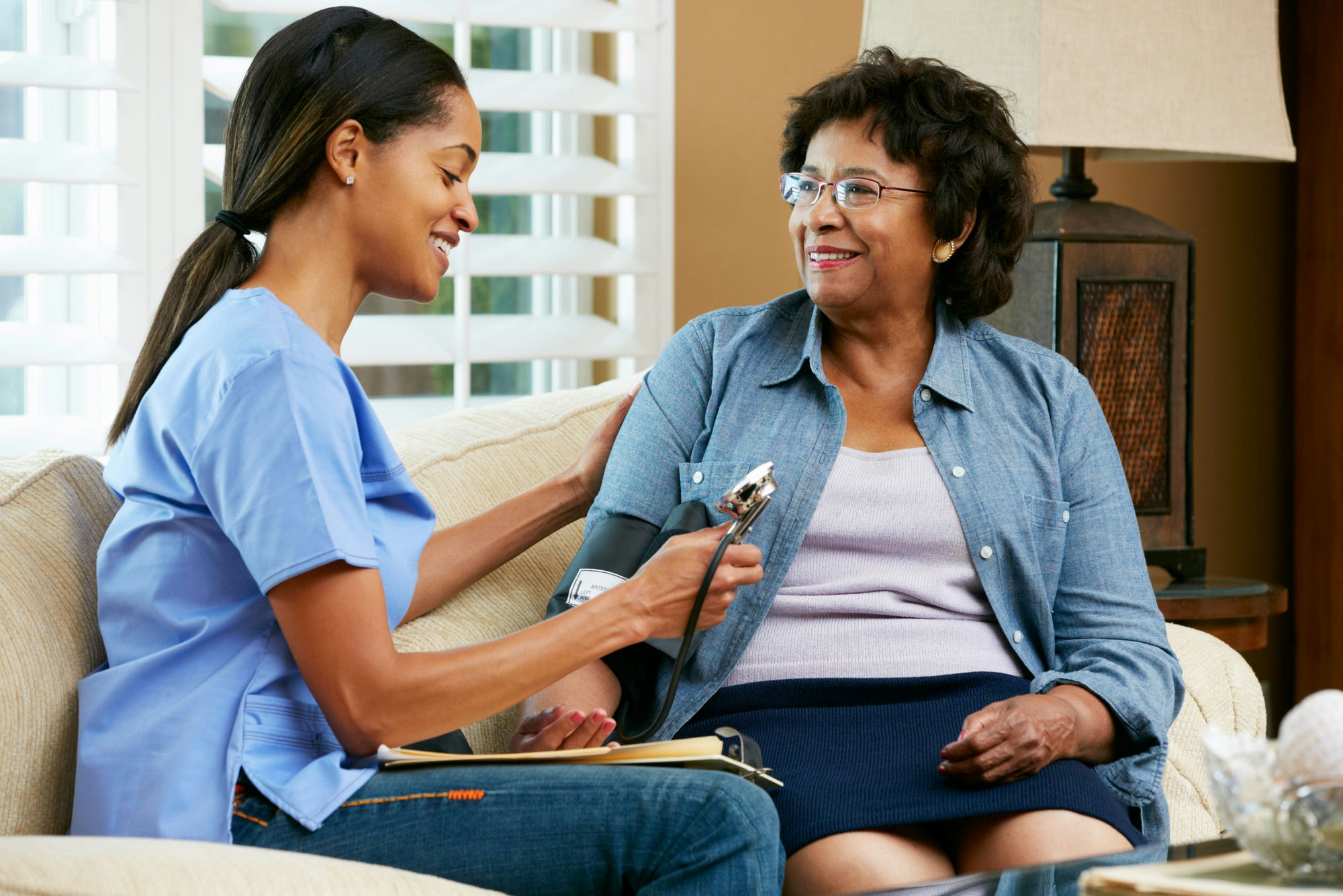 Caregiver in blue scrubs checking blood pressure of senior woman at home on couch — CareHub home care
